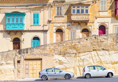 View of a typical maltese street with houses in traditional architecture style with colorful doors, balconies, windows and parked cars.