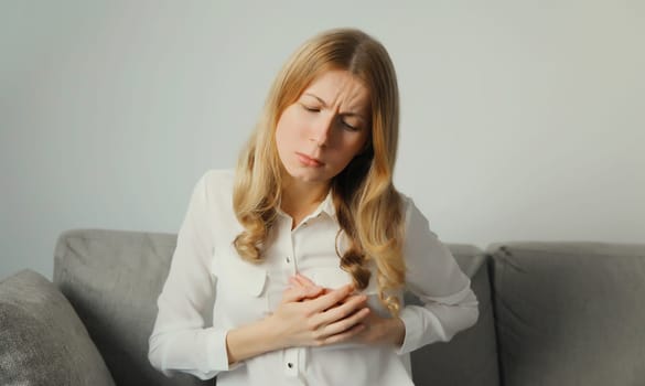 Unhealthy young woman touching hands her chest suffering from heart pain, feeling bad, heart attack, having health problems, sitting on the couch at home