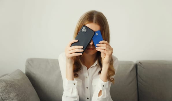Upset modern young woman holding smartphone and credit card sitting on the couch at home, internet fraud, problems with online payments or blocked bank card