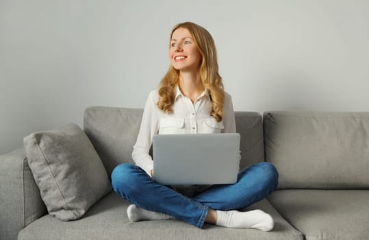 Happy modern young woman working with laptop, freelancer girl looks out the window using computer sitting on a couch in the living room at home