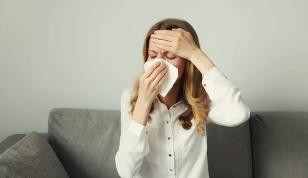 Sick exhausted young woman sneezing blow nose using tissue, unhealthy suffering girl feels unwell sitting on the couch at home