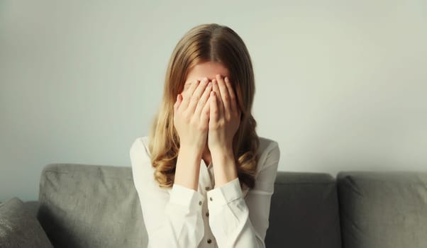 Upset sad crying woman covers her eyes and face with her hands while experiencing depression, stress, fatigue, mental suffering sitting on the couch at home