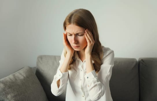 Exhausted tired woman touching head, suffering from headache, stress, fatigue, temples pain, migraine, loud noise, tension sitting on the couch at home