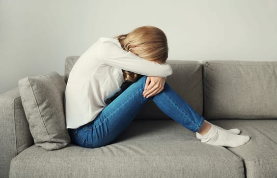Upset sad crying young woman covering her eyes and face with her hands while experiencing depression, stress, fatigue, mental suffering sitting on the couch at home