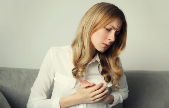 Unhealthy young woman touching hands her chest suffering from heart pain, feeling bad, heart attack, having health problems, sitting on the couch at home