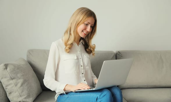 Happy modern young woman working with laptop, freelancer girl smiles looking at computer sitting on a couch in the living room at home