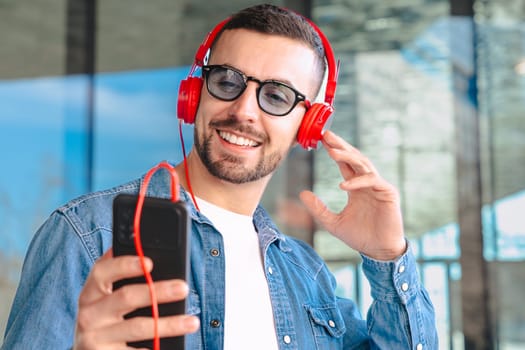 Smiling Caucasian man enjoys music through red headphones while holding a smartphone. Dressed in denim shirt, standing outdoors with modern glass building in background, bright and casual moment.