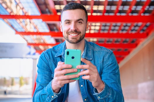 Smiling man in denim jacket holding smartphone and looking directly at the camera. Portrait in modern urban setting with red industrial beams and strong daylight. Stylish and focused expression.