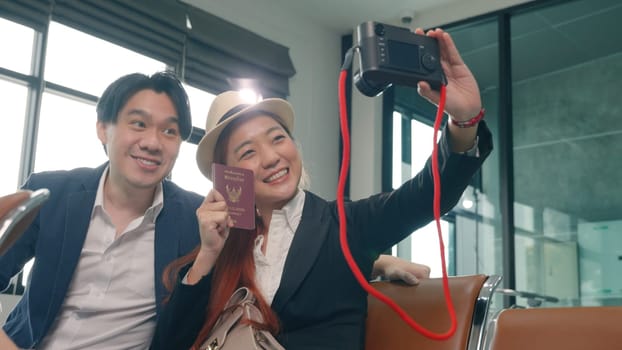 Wait boarding flight. Asian beautiful young loving couple smiling making selfie by their mirrorless camera and holding passports tickets while waiting for boarding in terminal hall airport
