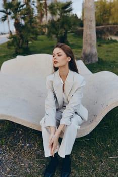 Fashionable woman wearing a light gray pantsuit sits thoughtfully on a stylish outdoor bench with lush green trees in spring, showcasing modern business style and calm mood