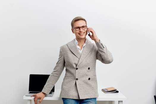 Smiling man wearing smart casual blazer and shirt standing in bright office with minimal decor, engaging in a phone conversation, showcasing a positive and professional attitude, with workspace items on desk, concept of business communication and success