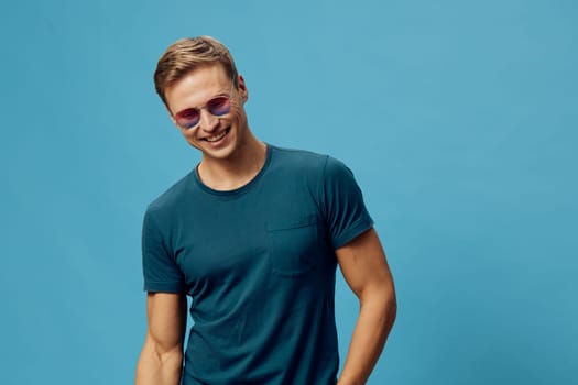 Smiling young man wearing casual dark teal t-shirt and purple sunglasses standing against blue background. Positive cheerful emotion, relaxed pose, fresh style. Concept: mental health and gadgets.