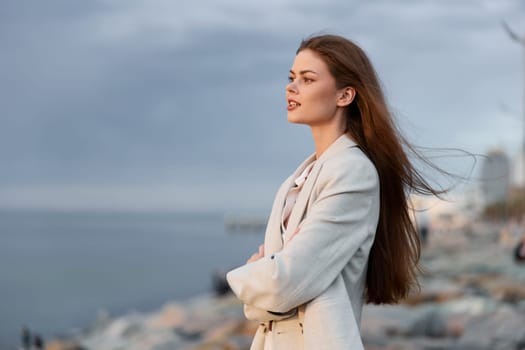 Thoughtful woman in beige coat with long hair stands by rocky seaside under cloudy sky, expressing calm and contemplation, autumn fashion and seasonal emotion by the water