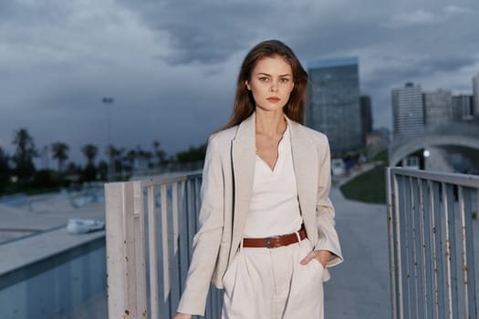 Confident young woman in beige business suit stands outdoors with urban skyline and cloudy sky background, showcasing professional style and serious emotion, suitable for business concepts.