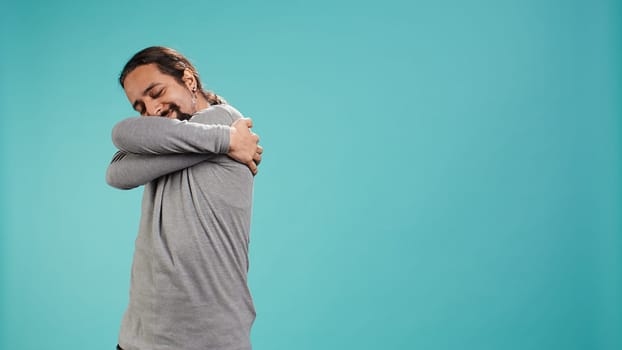 Portrait of man standing in studio, hugging himself in gesture of comfort and self love, isolated over studio background. Cheerful person doing self acceptance, embracing himself, camera B