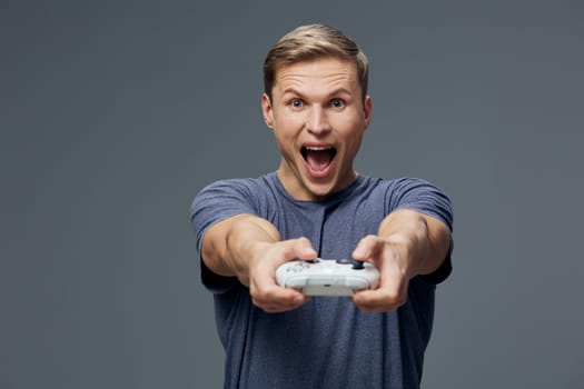 Joyful man in casual attire expressing triumph while playing video games with game controller in hands, smiling broadly, standing against neutral gray background, focused on entertainment and gaming lifestyle, concept of fun and relaxation