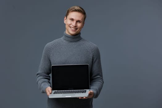 smiling young caucasian man holding laptop wearing gray turtleneck sweater standing isolated on gray background digital nomad working remote freelance business technology concept