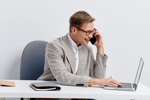 smiling young caucasian man wearing glasses and casual jacket talking on smartphone while typing on laptop at desk against plain white background. Businessman multitasking using digital devices for remote work. Concept: digital nomad lifestyle