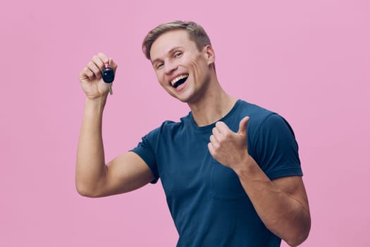 Happy man with short brown hair wearing casual blue t-shirt, smiling broadly while holding a car key, showing thumbs up gesture, isolated on pink background, conveying success, excitement, and achievement, lifestyle and optimism theme.