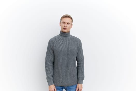 Studio shot of a young man with blonde hair, dressed in a cozy gray knitted turtleneck sweater, looking confidently at the camera, plain white background, casual style, fashion portrait, men lifestyle concept