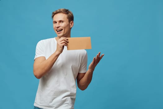 Bright studio shot of a smiling young man with short hair, dressed casually in a white t-shirt, holding a brown paper gift box. Blue background, joyful mood, celebrating, expressing excitement, lifestyle concept, happiness.