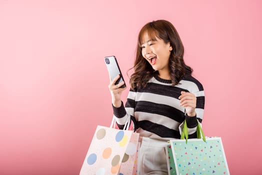 Portrait Asian happy beautiful young woman shopper smiling standing excited holding online shopping bags online multicolor and smartphone on hand, isolated on pink background, glad with mobile phone
