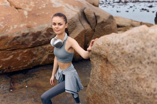 Fit young female model in sportswear and headphones navigating rocky landscape by the ocean, expressing focus and vitality amidst nature