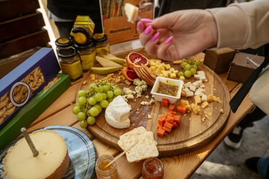 Cheese tasting at a farmers market, close-up of a woman's hand of a buyer. Cheese and fruit platter on the table. Cheese tasting process. Large assortment of natural products on the table. Hand in blur