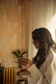 young romantic brunette with long wavy hair in a room with a cup and saucer in her hands. dressed in a white silk suit. stands in profile to the camera.
