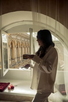 young brunette with long wavy hair stands in front of the window with a cup of coffee. she is dressed in a white silk suit. stands with her back to the camera.