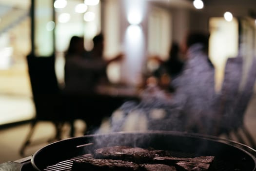 Close-up photo of delicious meat being grilled. In the background, friends and family are sitting and waiting for a meal