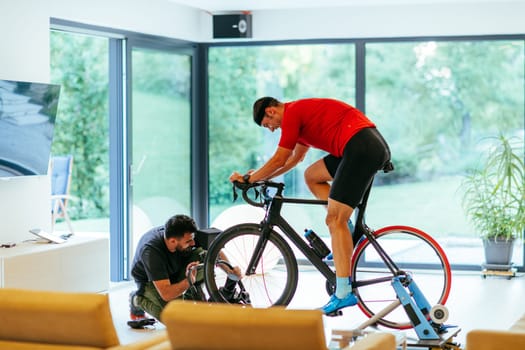 A cameraman filming an athlete riding a triathlon bike on a simulation machine in a modern living room. Training in pandemic conditions