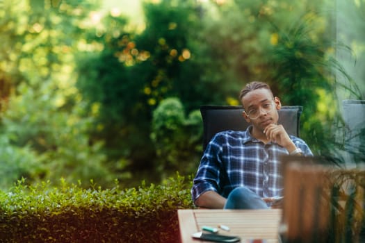 An African American man sitting in front of the house on a modern terrace and stares thoughtfully at the camera