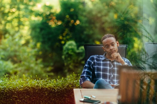 An African American man sitting in front of the house on a modern terrace and stares thoughtfully at the camera