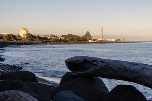 New Plymouth coastline view along Coastal Walkway beyond rocky foreshore and driftwood logs in foreground silhouette at sunrise looking down to port