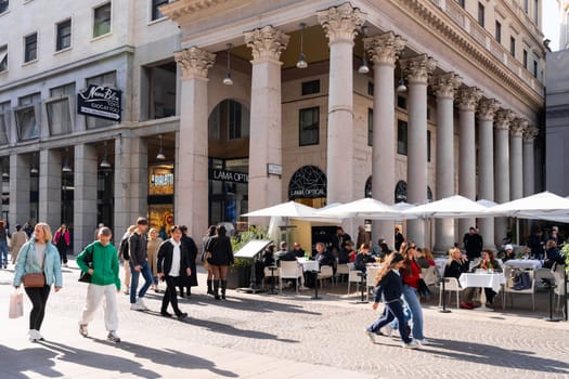 Milan, Italy - April, 08, 2025: Lama Optical and Bialetti storefronts under classic colonnade with people dining at cafe and walking by on sunny day in Milan.