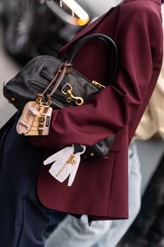Paris, France - March 11, 2025: Woman wears Miu Miu handbag in black leather with gold rings and mini charm keychains. Worn with wine-red blazer. Street style outfit details.