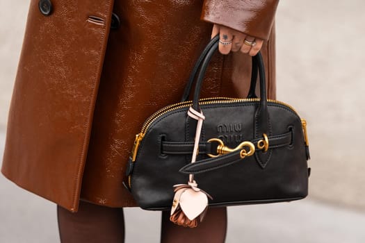 Paris, France - March 11, 2025: Black Miu Miu bag with gold details and charm. Brown high-shine patent trench coat. Street style outfit details.