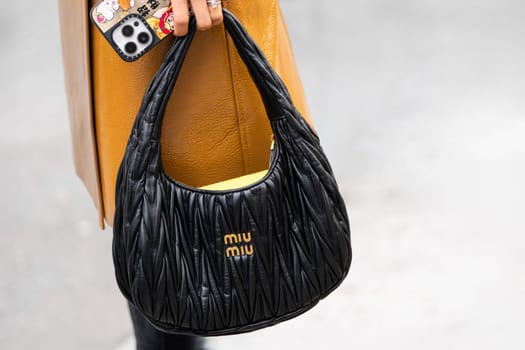 Paris, France - March 11, 2025: Woman wears black ruched leather Miu Miu bag with gold logo, paired with tan skirt. Street style outfit details.