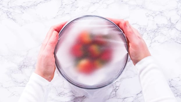 Flat lay. Strawberries, freshly washed and dried, are neatly stored in a glass bowl lined with a paper towel and securely covered with plastic wrap to maintain freshness.