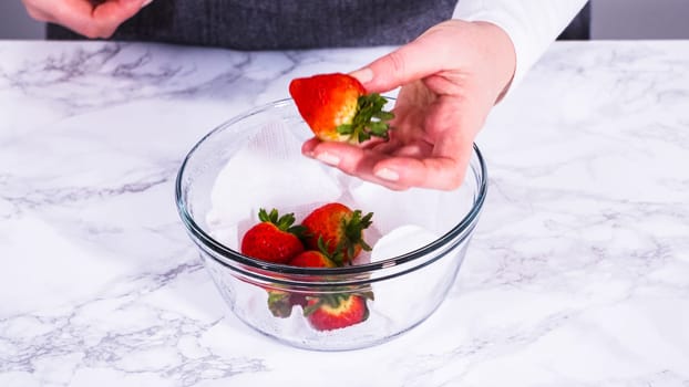 Strawberries, freshly washed and dried, are neatly stored in a glass bowl lined with a paper towel and securely covered with plastic wrap to maintain freshness.