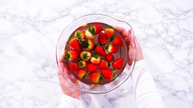 Flat lay. Ripe strawberries are submerged in water within a large glass mixing bowl, a step in washing the fruit to ensure cleanliness and longevity before storage or consumption.