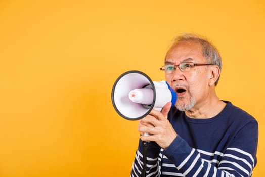 Portrait Asian old man wearing glasses screaming in megaphone studio shot isolated yellow background, happy elderly man holding a megaphone and shouts yelling announces news information