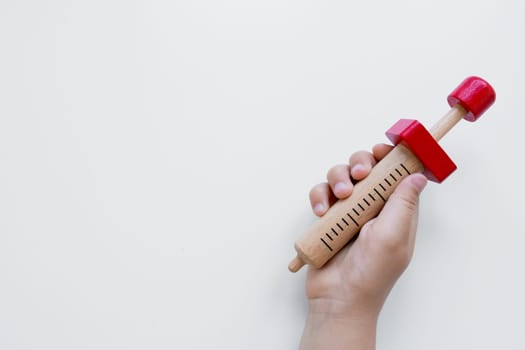 Child hand holding wooden toy syringe with red accents on white background.
