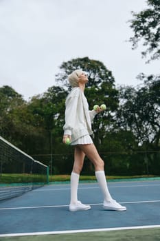 Energetic young female athlete in stylish sports attire holding weights on a tennis court, surrounded by greenery, conveying themes of health, fitness, and determination