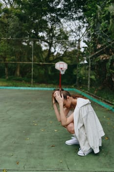 A young female athlete in a white hoodie and casual shorts expresses frustration on a lush outdoor basketball court, embodying emotional depth and resilience