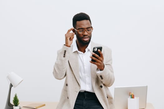 Confident young man wearing glasses and beige blazer using smartphone in minimalist office setting, focused on screen, working remotely or communicating, professional and stylish appearance, clean workspace.