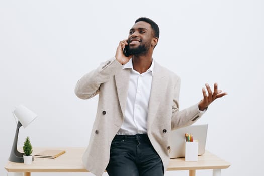 Cheerful man wearing beige blazer and white shirt engaging in a phone call, smiling broadly, standing beside a minimalist desk with office accessories, on a clean white backdrop. Professional and positive atmosphere.