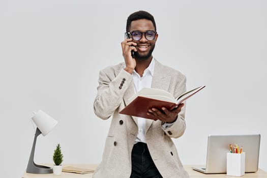 Confident smiling man with glasses dressed in beige blazer engaging in a phone conversation while holding an open book. Modern office background with a laptop and small plant, emphasizing professional environment. Concept: workplace communication.