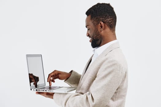 Confident man with dark skin, dressed in a light beige suit, engaging with a laptop computer, standing against a plain white background. Emphasizes technology, remote work, professionalism, modern business lifestyle, concentration, innovation.